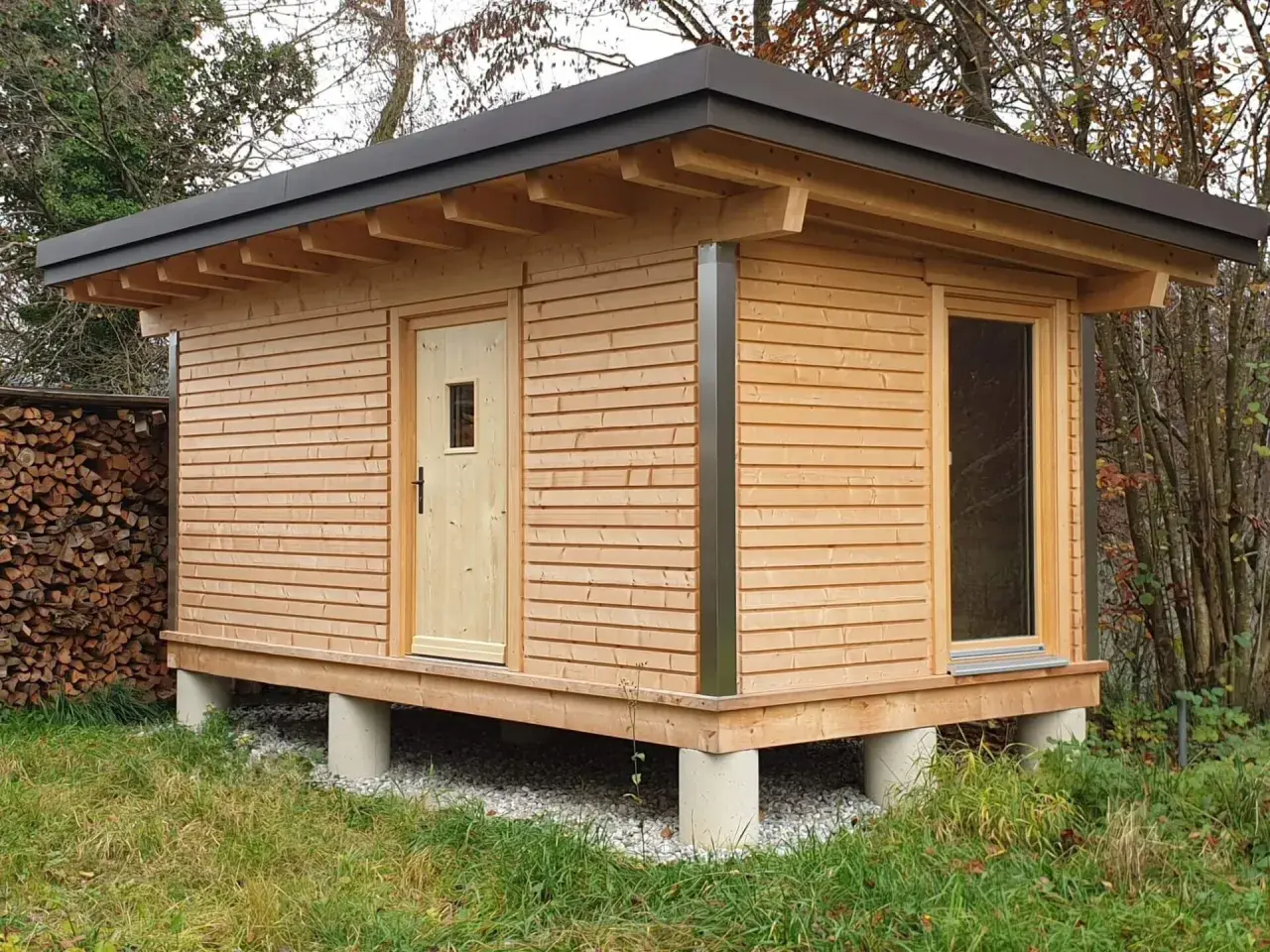 Moderne gartenhaus ideen: ein Holzhaus auf Betonpfeilern mit Pultdach, Holztür und großem Fenster. Daneben ein Holzstapel.