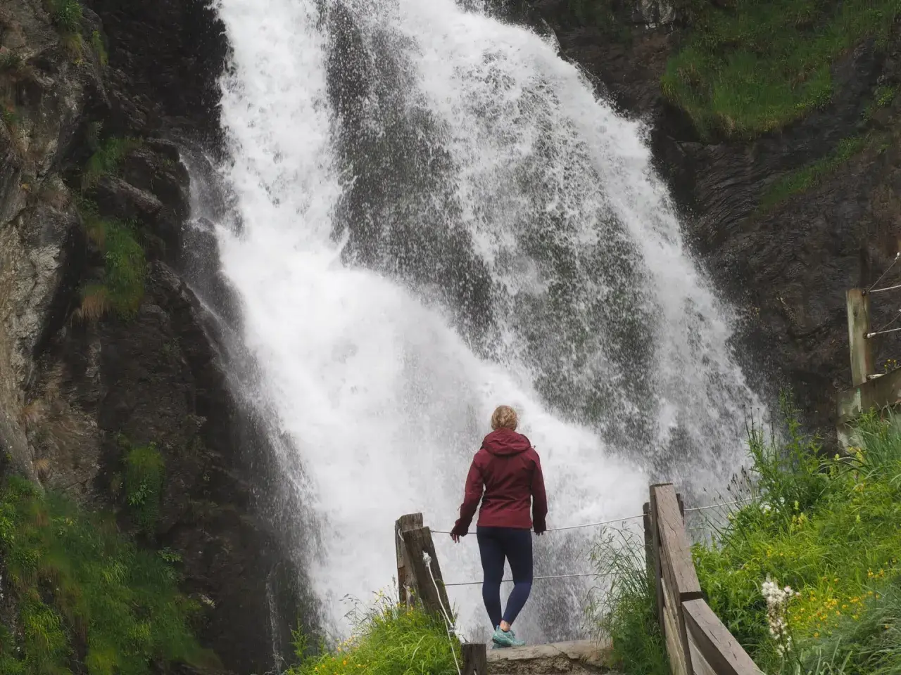 Mujer contemplando una cascada en Vielha e Mijaran. El agua cae con fuerza sobre rocas oscuras, rodeada de vegetación verde.