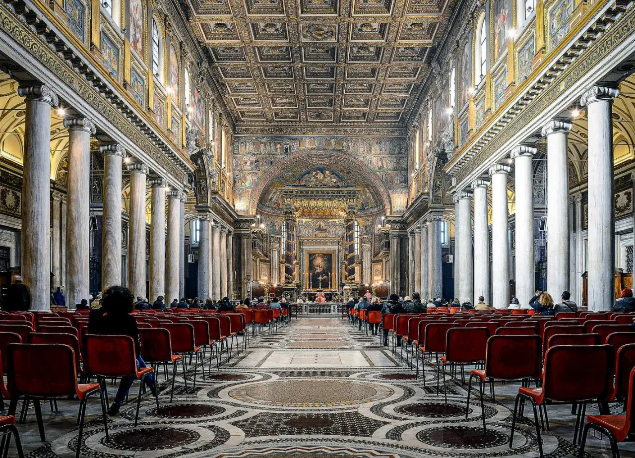 Intérieur de Sainte-Marie-Majeure, Rome. Des fidèles assis sur des chaises rouges, sous des colonnes imposantes et un plafond orné.