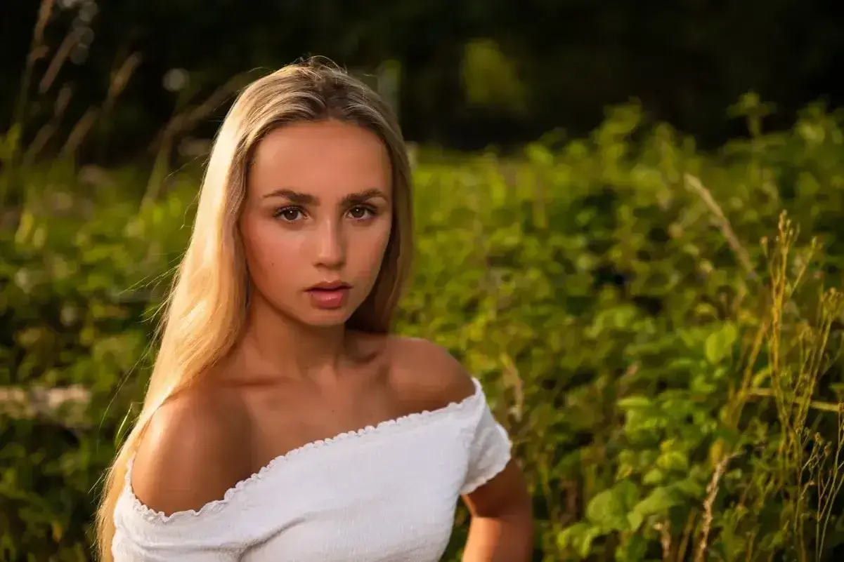 Młoda kobieta z długimi blond włosami, ubrana w białą, odkrywającą ramiona bluzkę, patrzy prosto w obiektyw na tle zielonej, rozmytej roślinności.