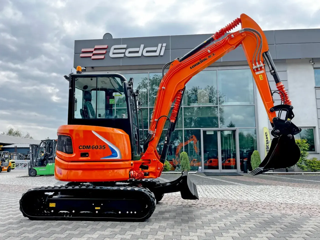 Pomarańczowa koparka gąsienicowa CDM 6035 przed budynkiem z logo Eddi.