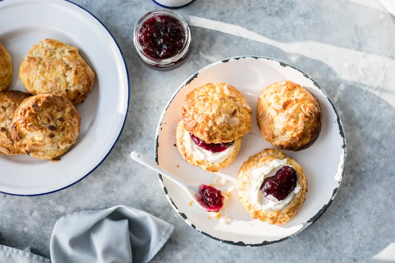 Nybakade scones med mandelflagor, grädde och sylt på en tallrik. En härlig fika!
