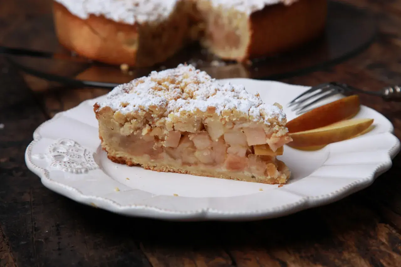 Ein Stück apfelkuchen sehr fein, mit Streuseln und Puderzucker bestäubt, auf einem weißen Teller mit Gabel und Apfelscheiben.