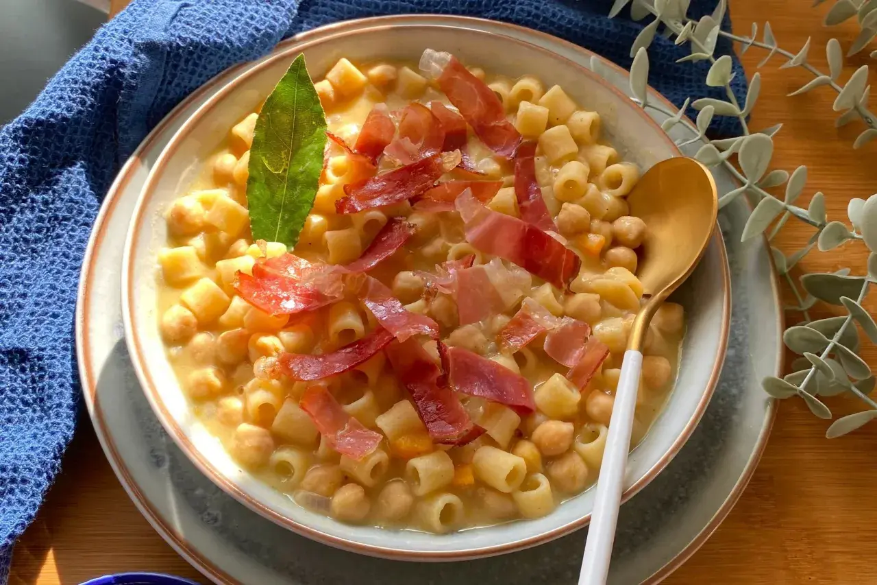 Un piatto fumante di pasta e ceci, arricchito da fette di prosciutto crudo e una foglia di alloro.