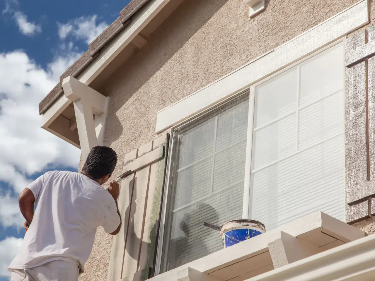 Pintor aplica pintura a persianas de ventana. ¿Cuál es la mejor pintura para fachadas? Busca durabilidad y resistencia a la intemperie.