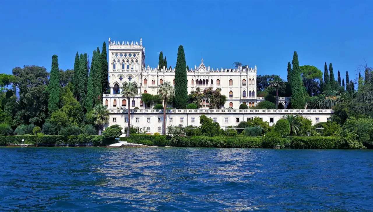 Villa neogotica su un'isola del Lago di Garda, circondata da cipressi e vegetazione lussureggiante, con il blu intenso dell'acqua in primo piano.