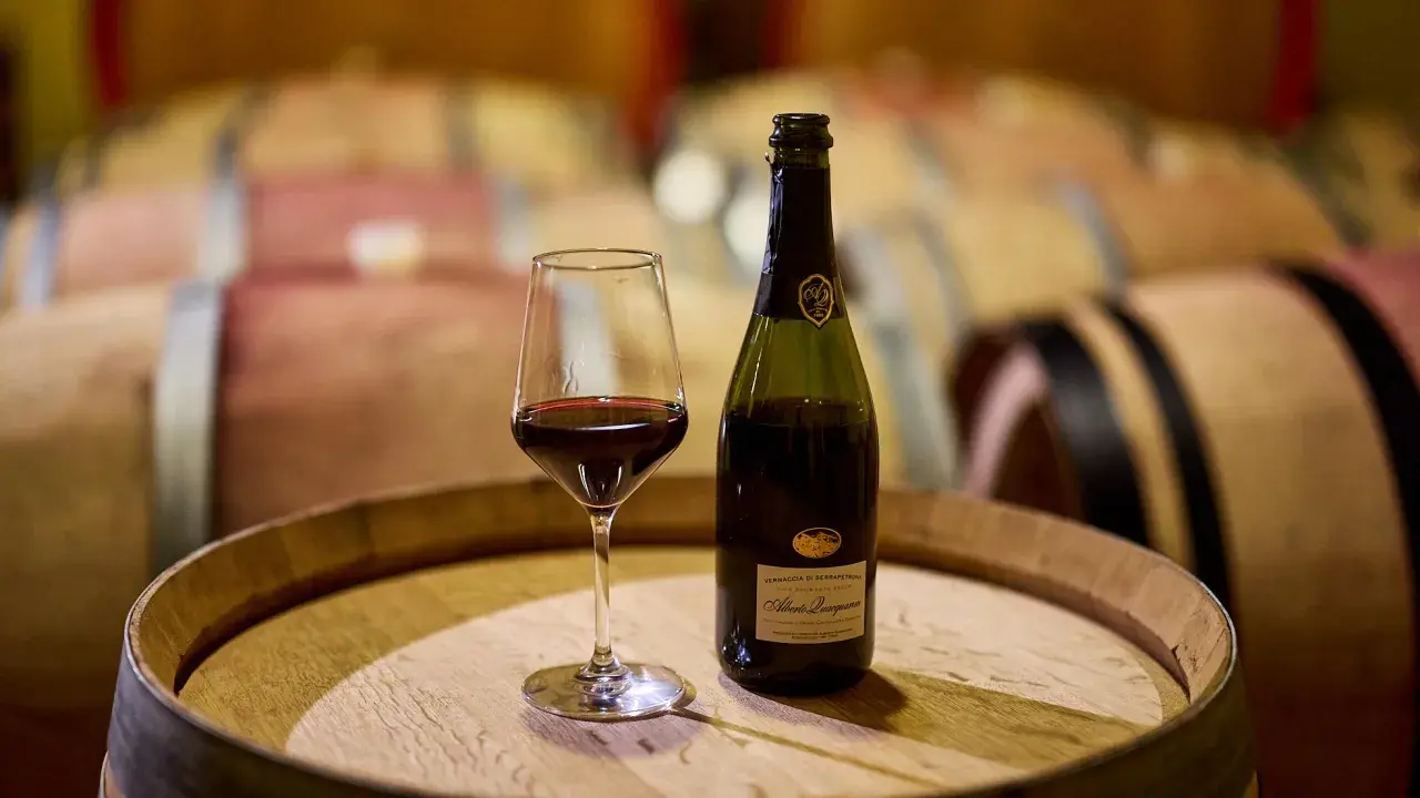 Calice di vernaccia serrapetrona e bottiglia su una botte di legno, con botti in cantina sullo sfondo.