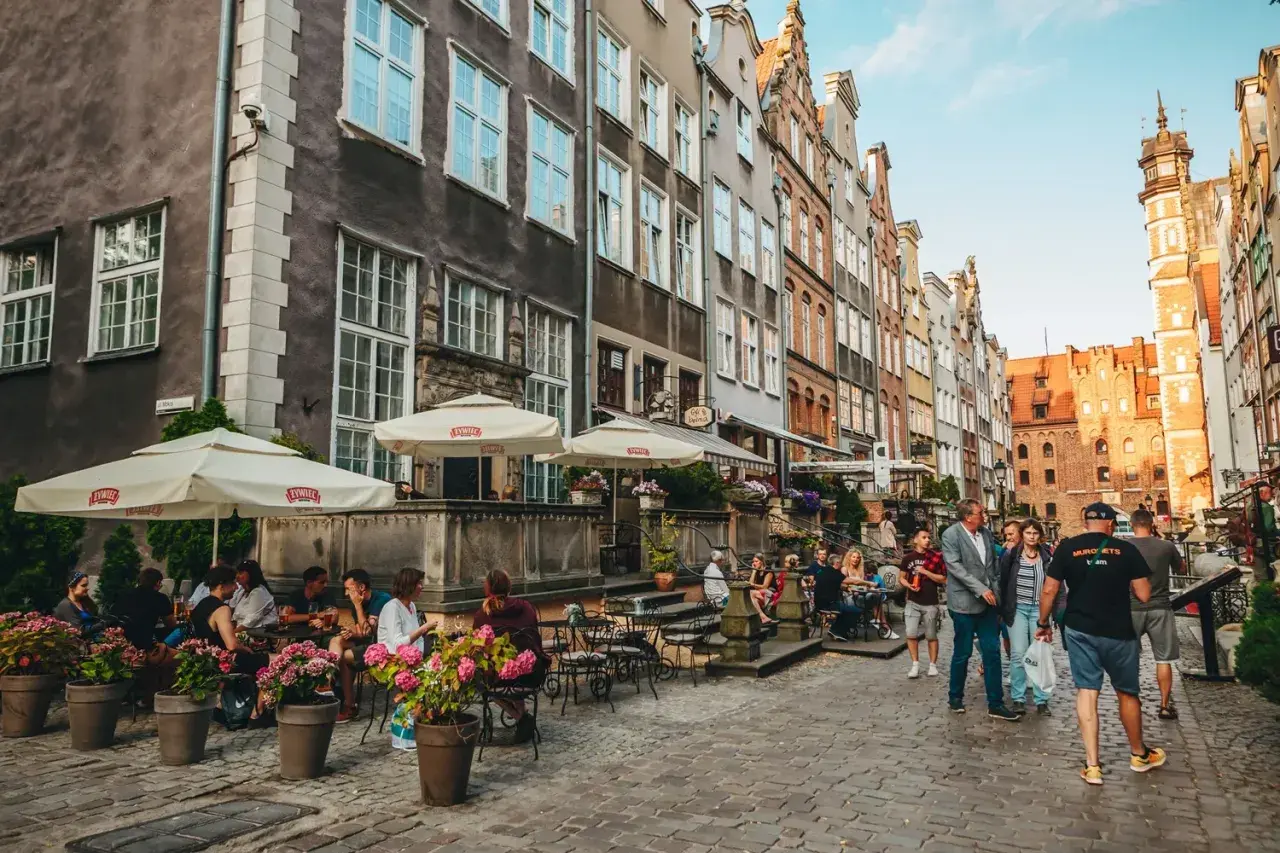 Ludzie siedzą przy stolikach pod parasolami, ciesząc się posiłkiem w jednej z urokliwych restauracji Gdańsk Starówka. Kamienne uliczki i zabytkowe kamienice tworzą niepowtarzalny klimat.