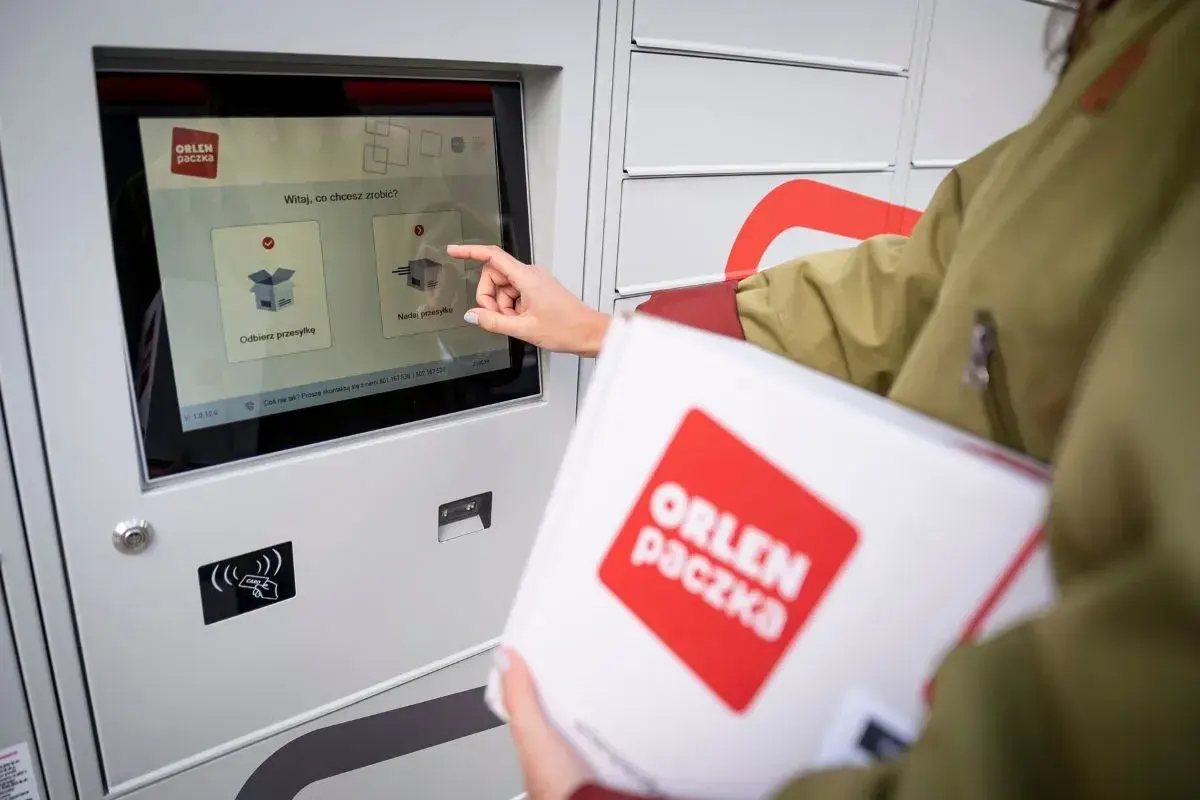 Dłoń dotyka ekranu automatu paczkowego, wybierając opcję "Nadaj przesyłkę". W ręku trzymana jest biała paczka z logo ORLEN paczka.