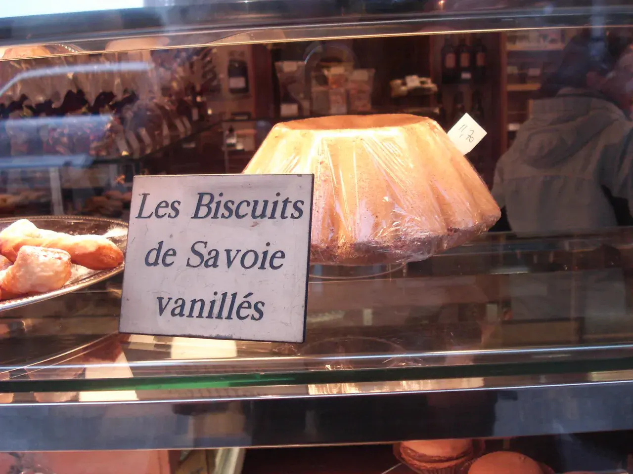 Un grand biscuit de Savoie vanillé, emballé sous plastique, est présenté dans une vitrine de boulangerie.