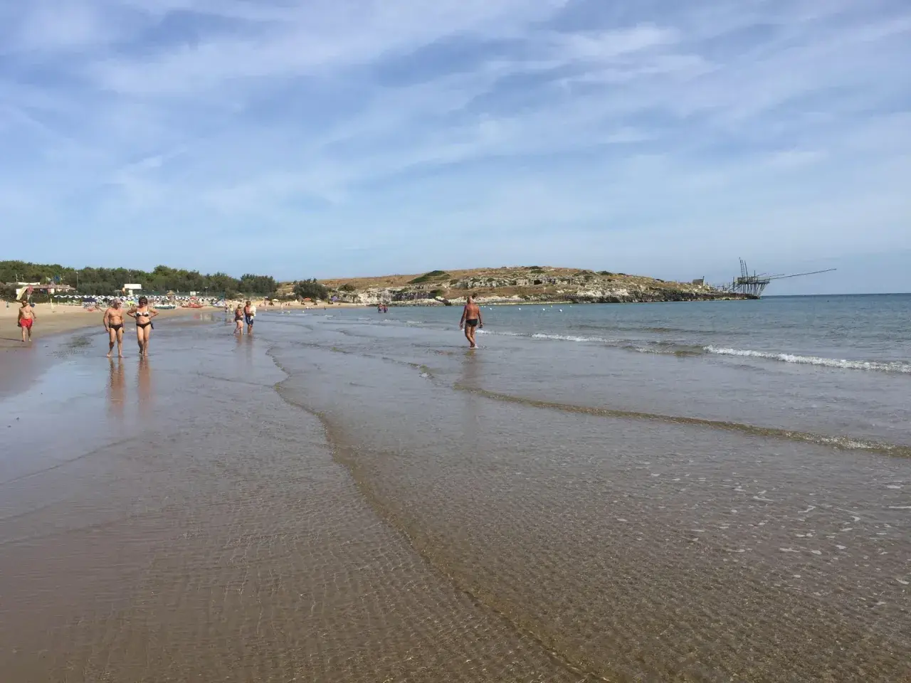 Persone passeggiano sulla battigia di Cala Molinella, con un trabucco sullo sfondo.