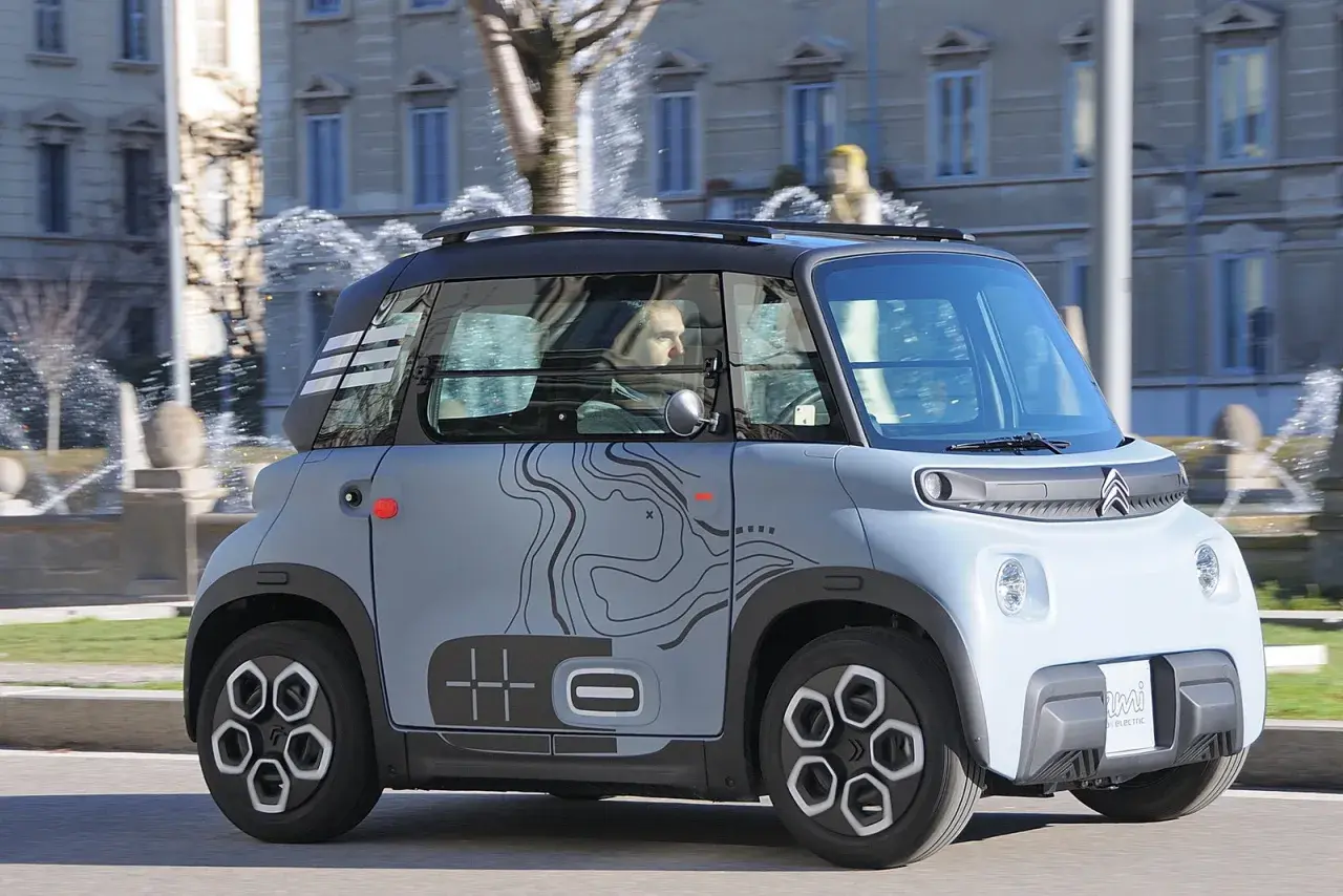 Citroën Ami, un veicolo elettrico compatto, sfreccia con la sua velocità massima in città, con uno sfondo di fontane e palazzi.