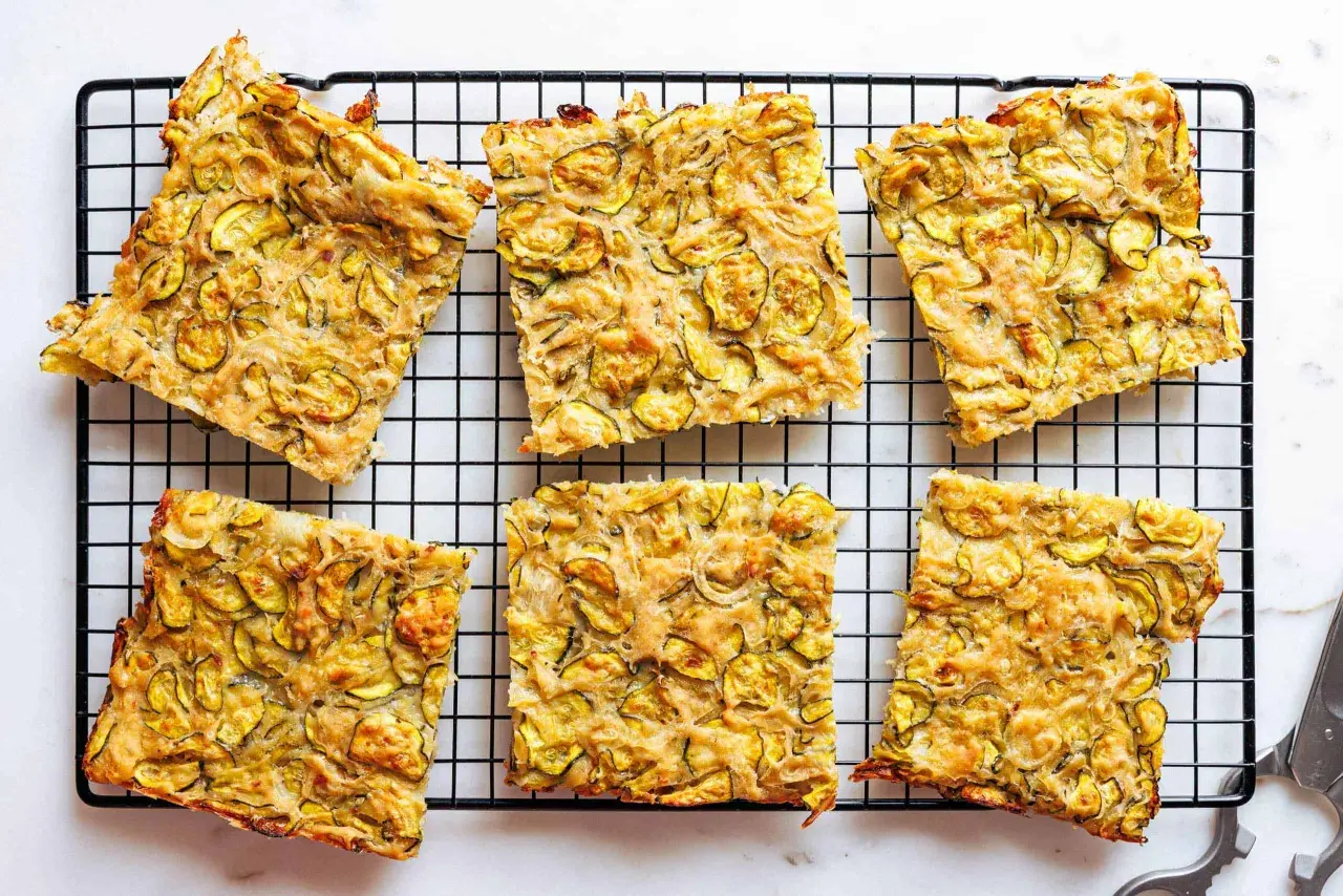 Sechs rechteckige Stücke einer gebackenen Zucchinipfanne mit gelben Zucchinischeiben auf einem schwarzen Gitter.