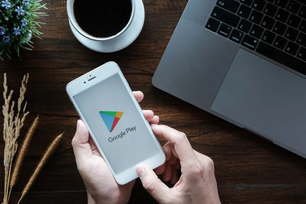 Ręce trzymają smartfon z logo Google Play, gotowe by dowiedzieć się, jak zaktualizować usługi Google Play. Obok laptop i kawa.