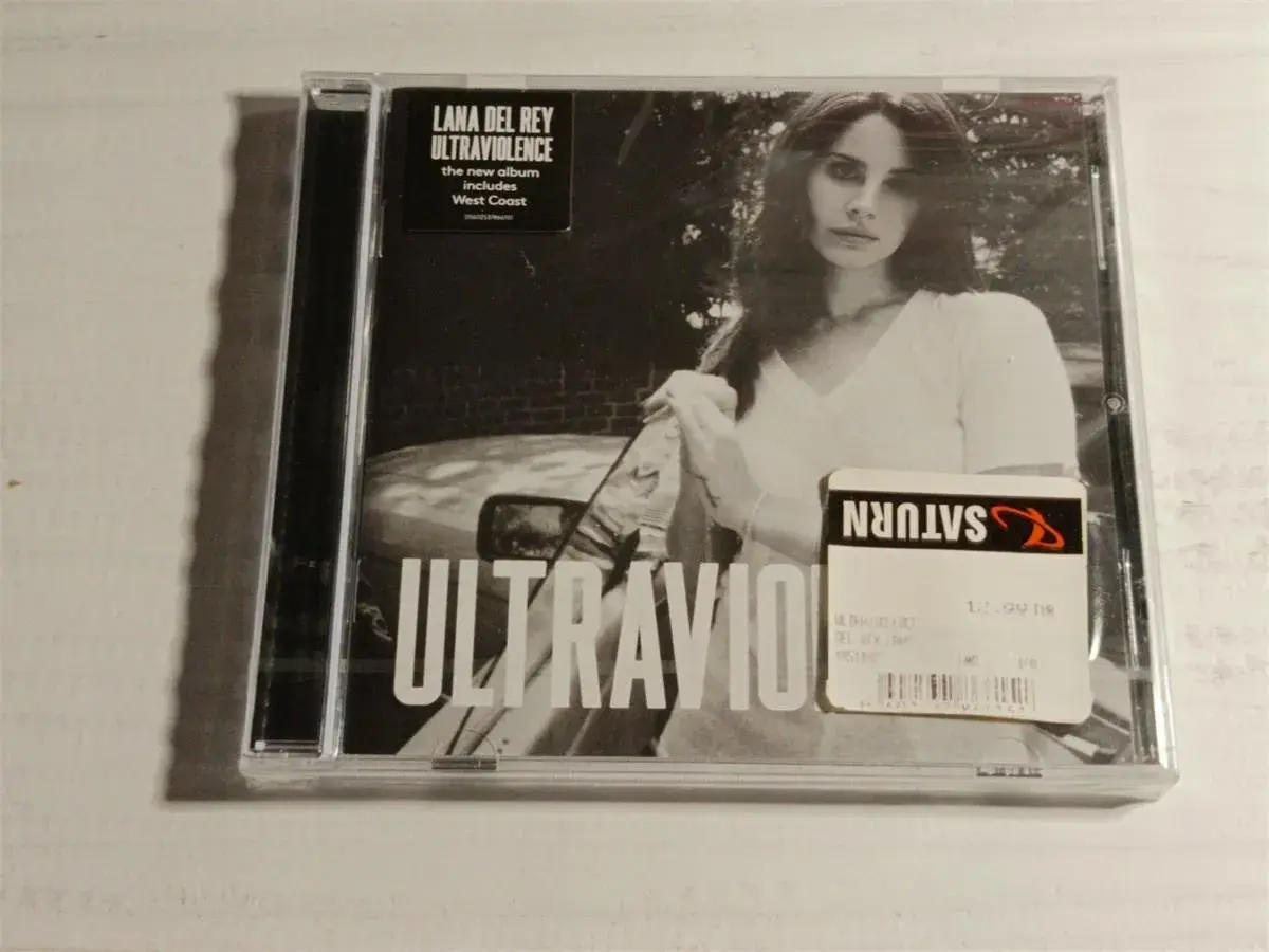 Okładka płyty Lana Del Rey Ultraviolence. Na czarno-białym zdjęciu artystka w białej koszulce, obok naklejka Saturn z ceną.