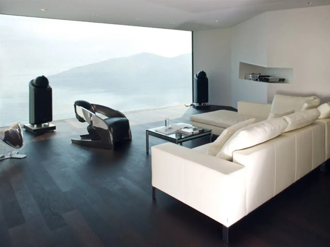 Modernes Wohnzimmer mit Panoramafenster, das eine atemberaubende Aussicht bietet. Ein weißes Ledersofa, ein futuristischer Sessel und zwei schlanke Lautsprecher ergänzen die minimalistische Einrichtung. Das Panoramafenster dekorieren wir hier mit der N...