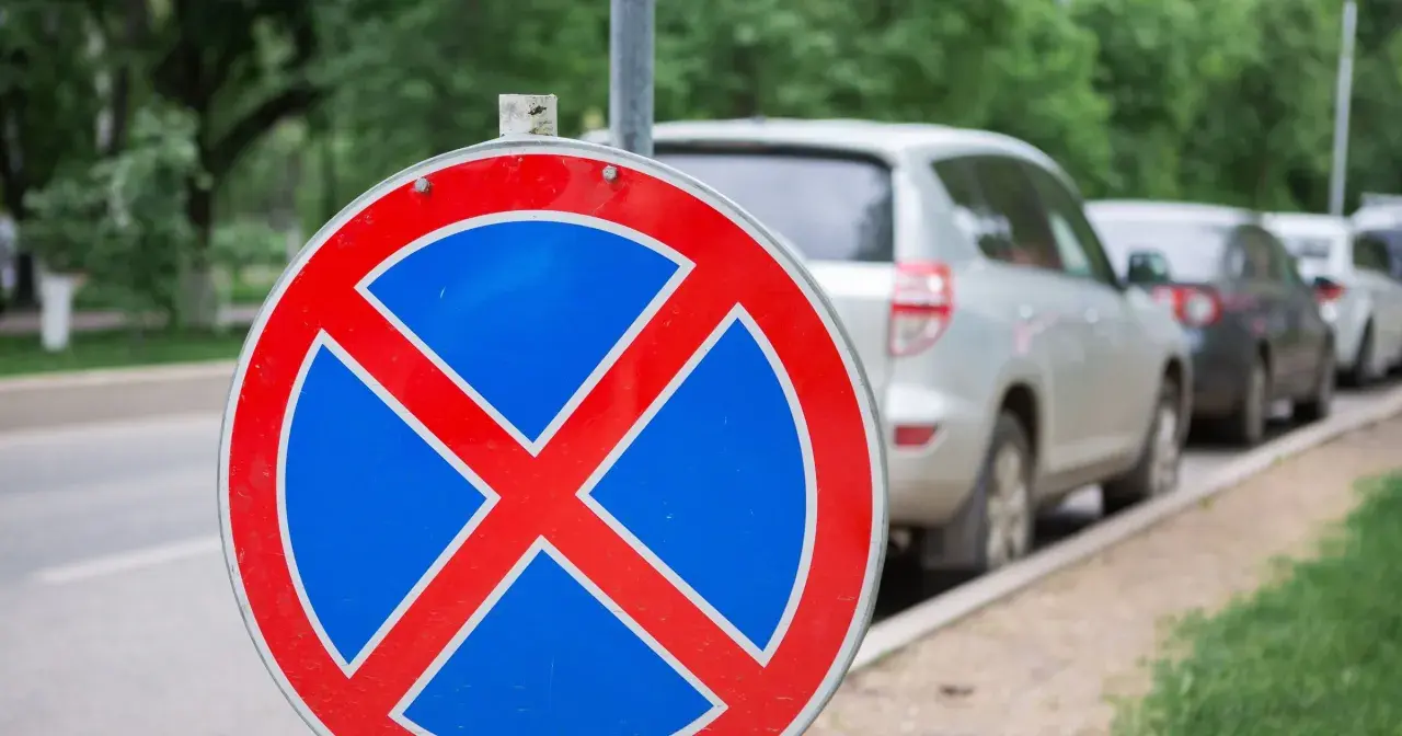 Znak zakazu zatrzymywania się b36. Samochody stoją w rzędzie przy ulicy, ale ten znak przypomina, że nie wolno tu parkować.