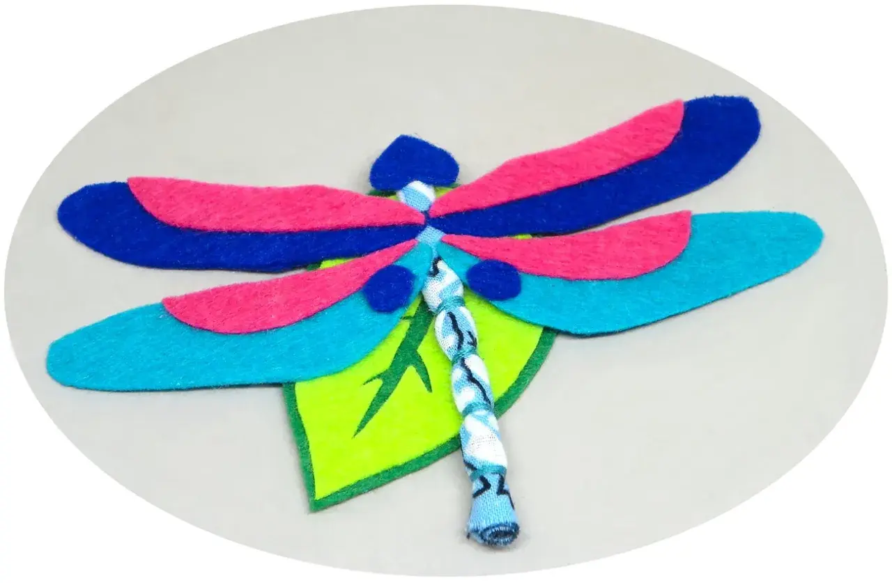 Libélula de fieltro, un proyecto de manualidades con alas azules, rosas y turquesas, posada sobre una hoja verde.