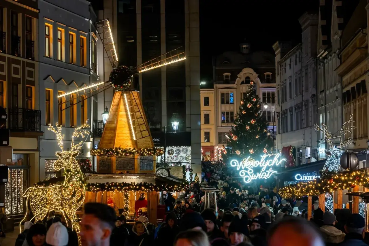 Tłum ludzi na jarmarku bożonarodzeniowym w Kujawsko-Pomorskiem. Świecący renifer, drewniany wiatrak i choinka tworzą magiczną atmosferę.