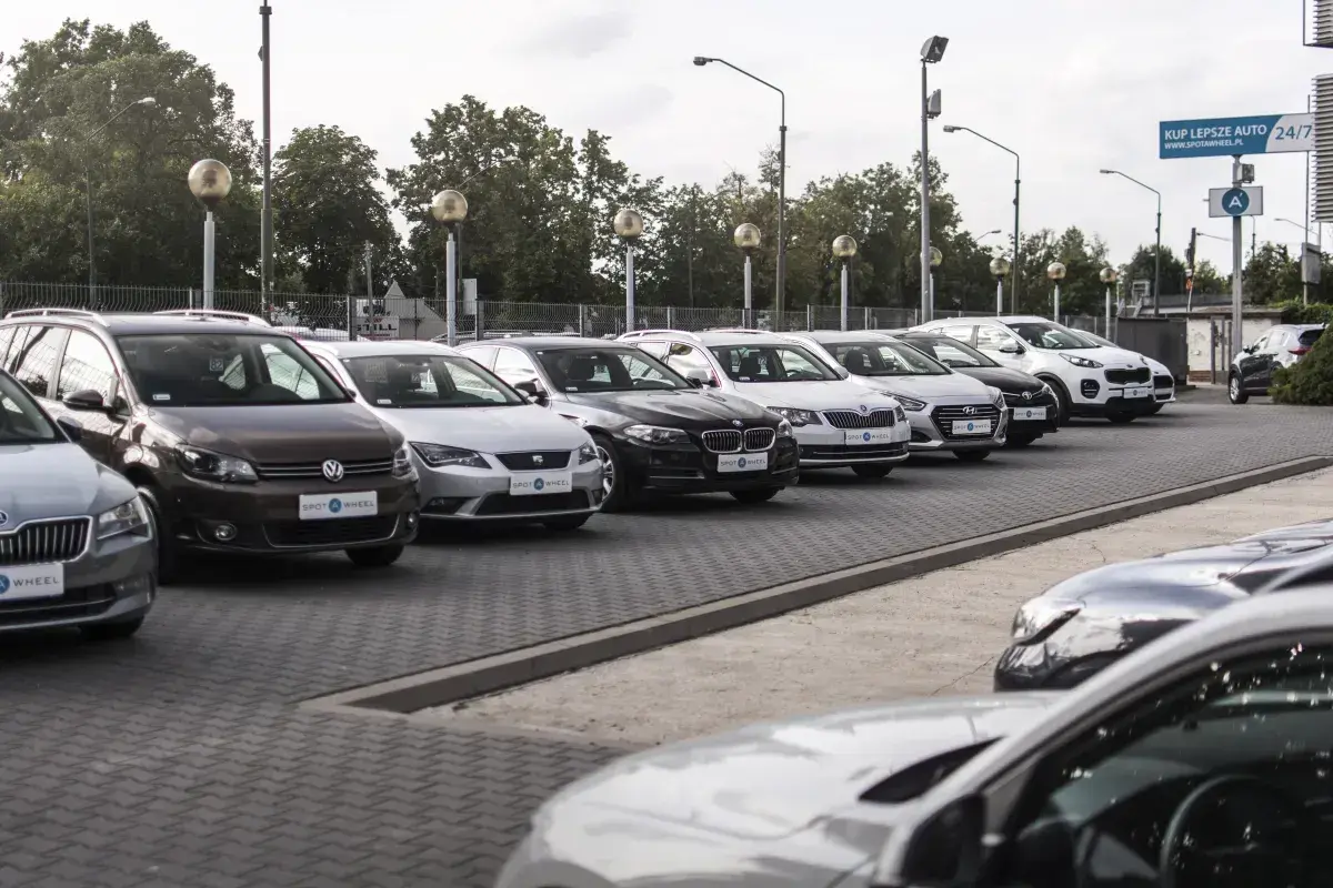 Rząd samochodów na placu, w tym brązowy Volkswagen i biały Kia Sportage. W tle drzewa i słupy oświetleniowe. To idealne miejsce, by znaleźć swój wymarzony kinto one.