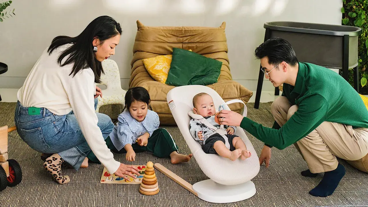 Familjen leker på golvet. Bebisen sitter i en Stokke babysitter medan pappan leker med den. Systern leker med en träleksak.