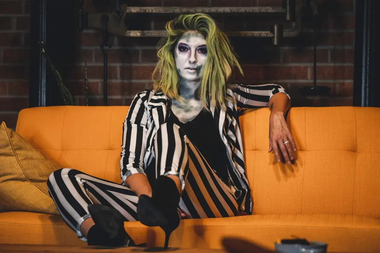 Donna con trucco spettrale e capelli verdi, indossa un vestito da strega fai da te a righe bianche e nere, seduta su un divano arancione.