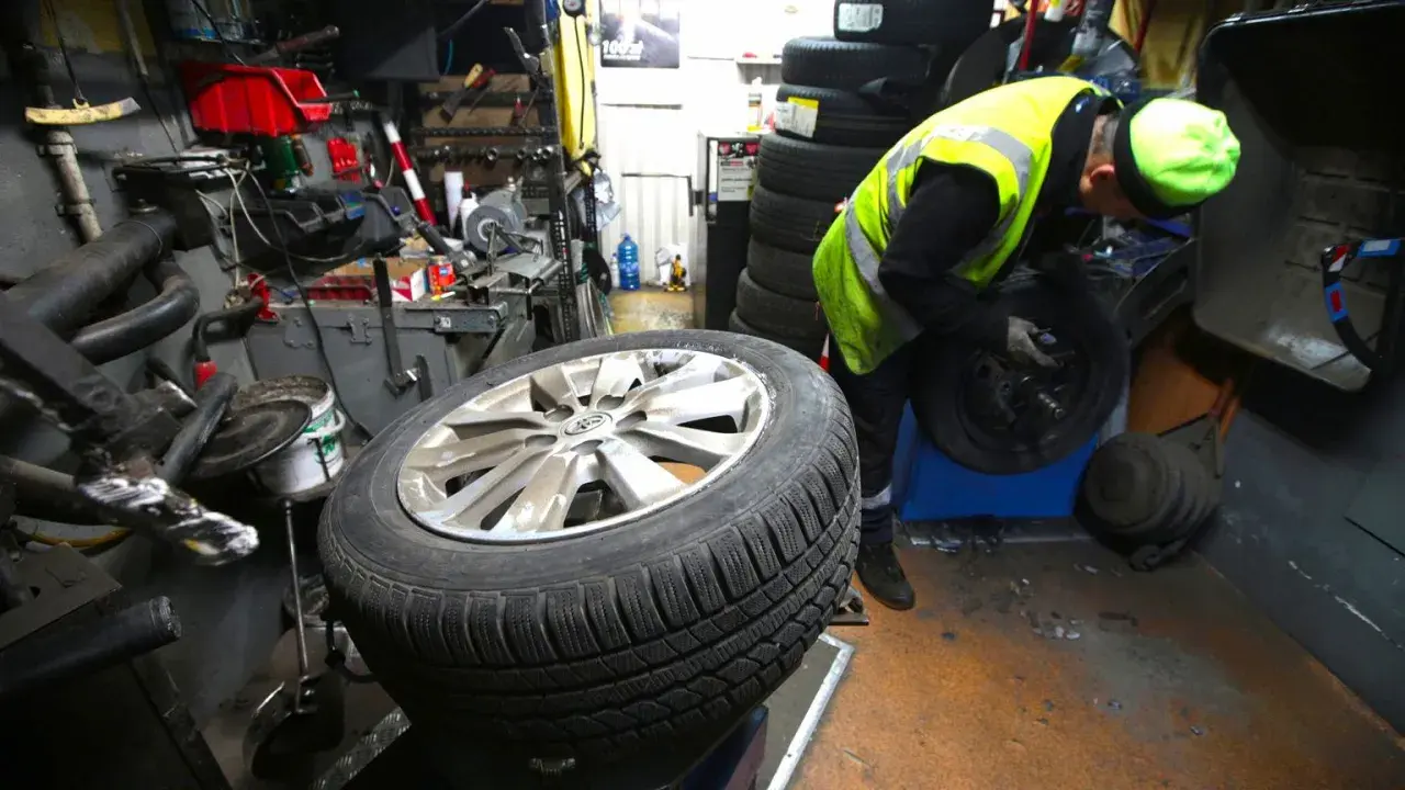 Mechanik w kamizelce odblaskowej pracuje przy kole. Słychać buczenie opon podczas jazdy, gdy pracownik wymienia oponę.