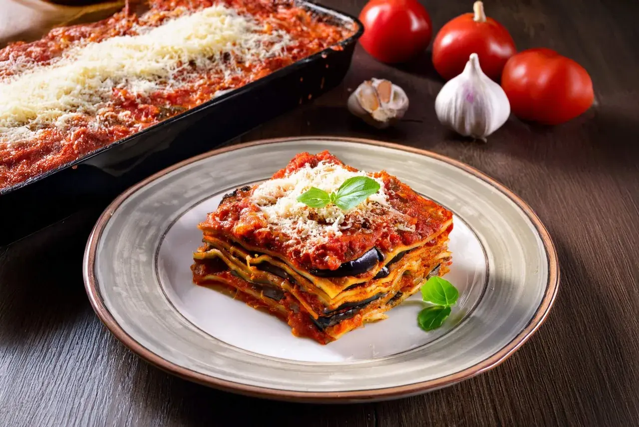 Una fetta di lasagne vegetariane, con melanzane e pomodoro, guarnita con basilico e parmigiano. Sullo sfondo, ingredienti freschi.