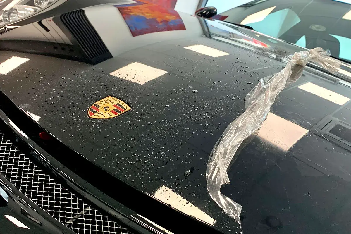 Czarna maska Porsche z logo, na której widać krople wody i fragment folii. Trwa klejenie folii na sucho.