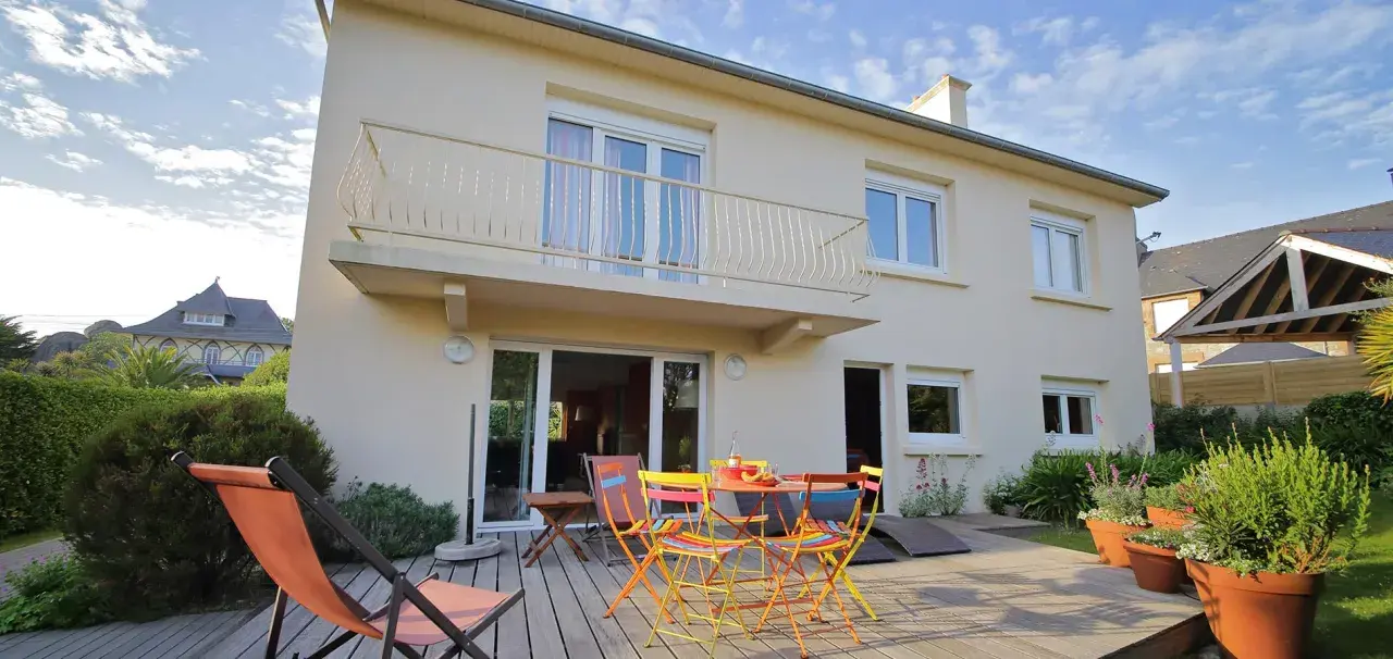 Maison divisée en 2 logements avec terrasse en bois, mobilier coloré et transat. Jardin verdoyant et ciel bleu.