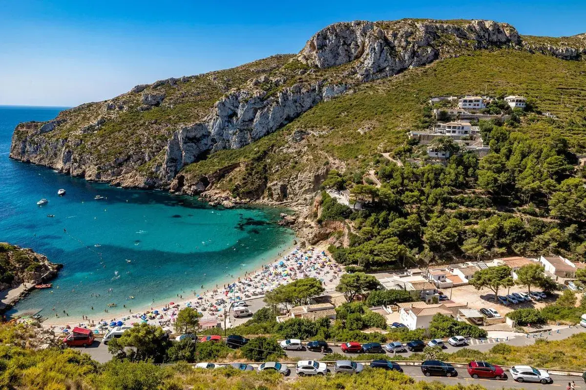 Bahía con aguas turquesas y playa abarrotada, rodeada de acantilados y vegetación. Un paraíso entre las mejores playas de Alicante.
