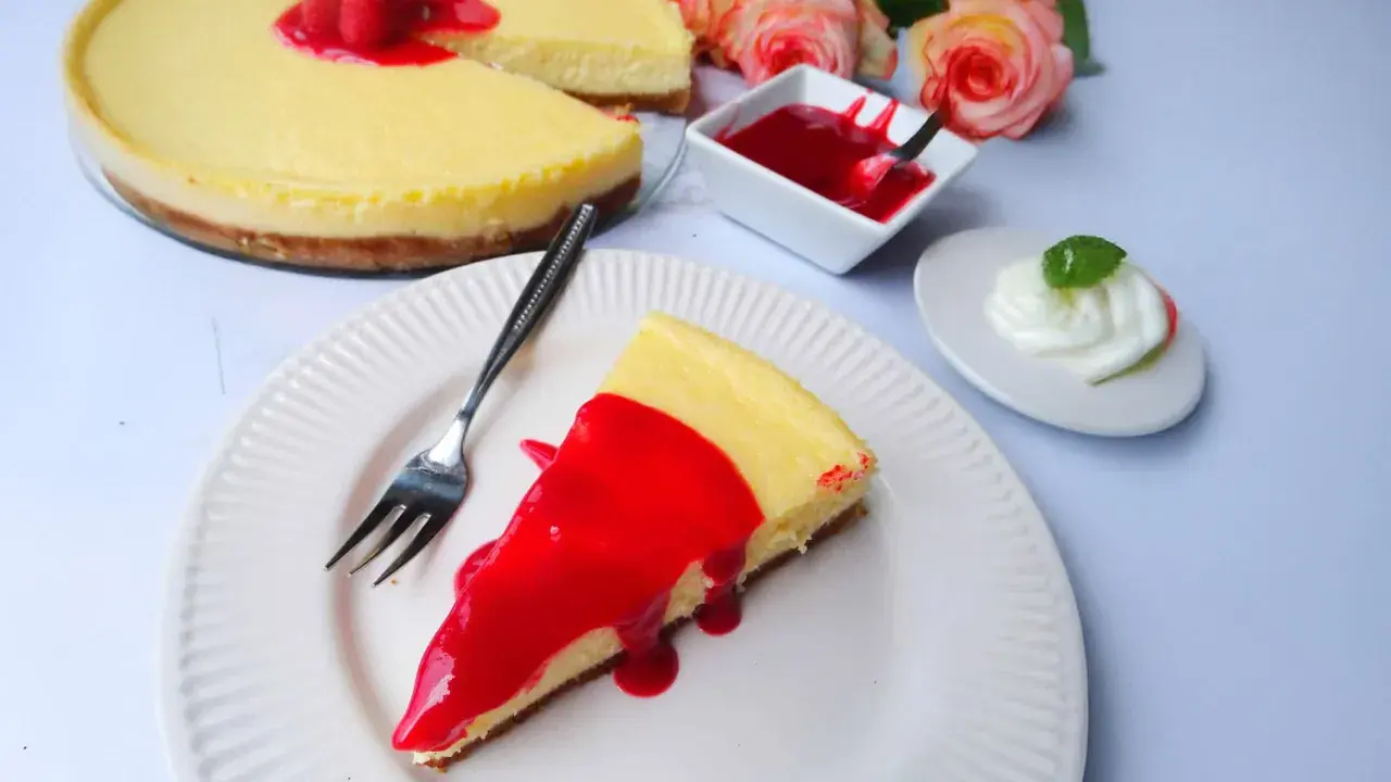 Ein Stück New York Cheesecake mit roter Fruchtsauce und Himbeeren, daneben eine kleine Schüssel Sauce und ein Klecks Sahne.