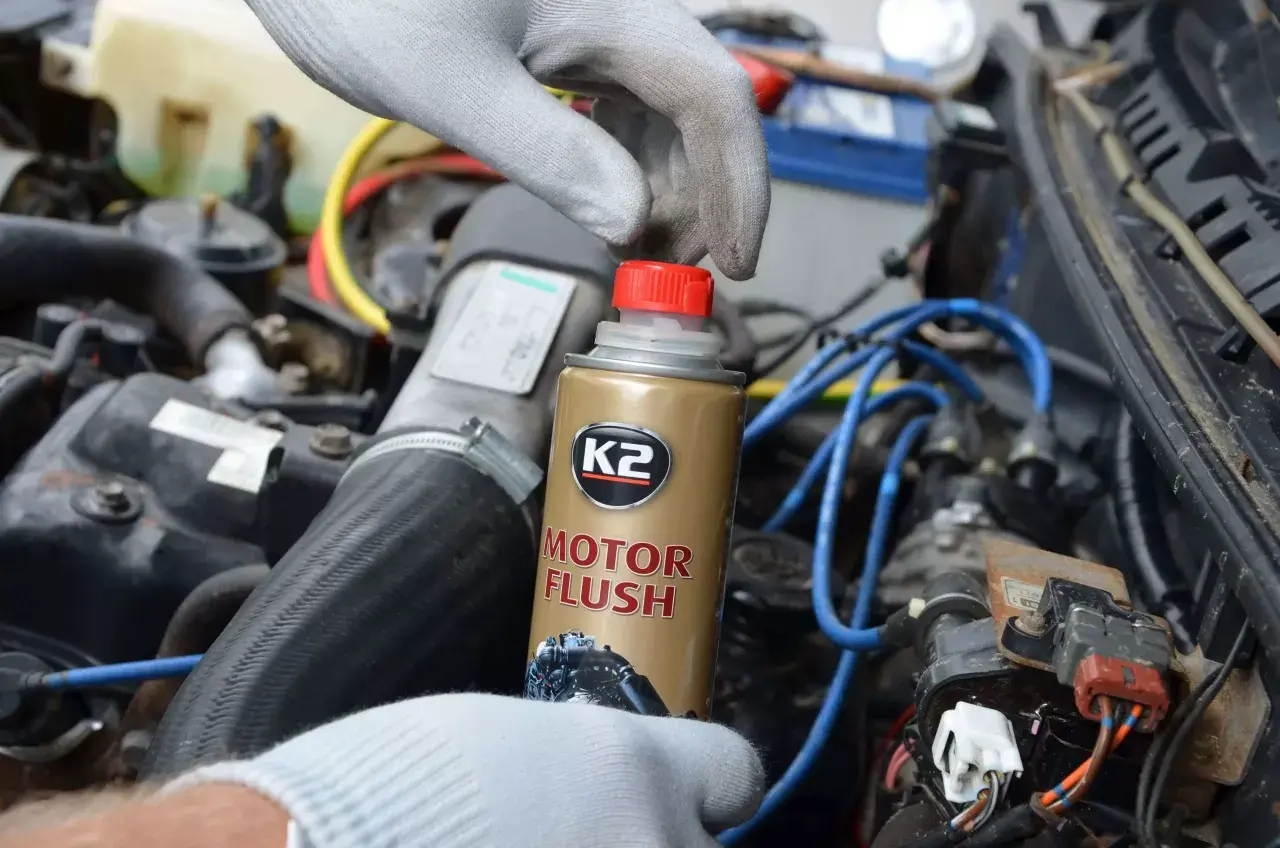 Ręka w rękawiczce trzyma puszkę płukanki do silnika K2 Motor Flush na tle silnika samochodowego.