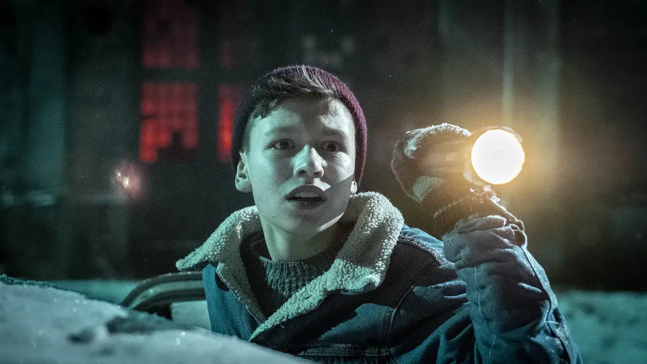 Chłopiec w zimowej kurtce i czapce trzymający latarkę, oświetla mroczny, ośnieżony teren. Może szuka czegoś w pobliżu czarnego młyna.