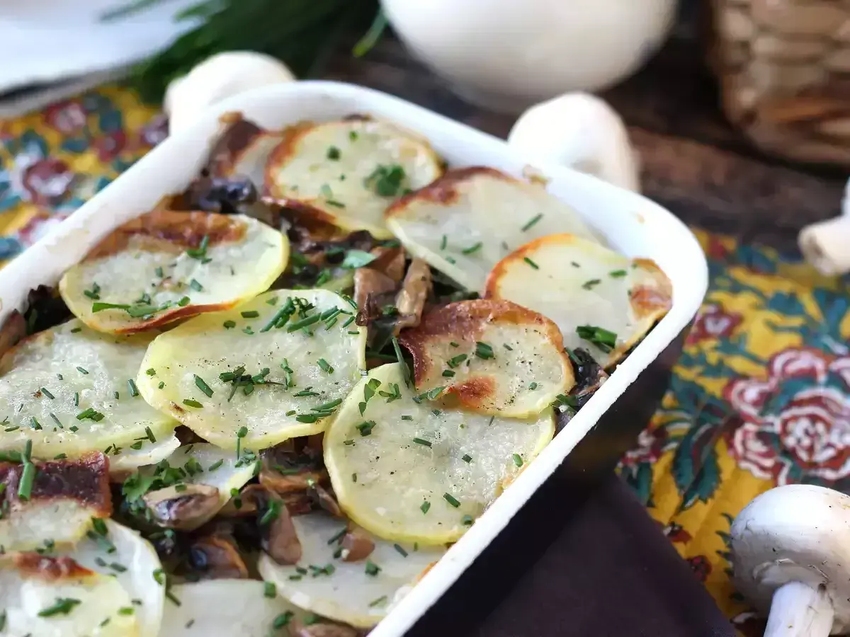 Zapiekanka ziemniaczana z pieczarkami, posypana szczypiorkiem, w białym naczyniu.