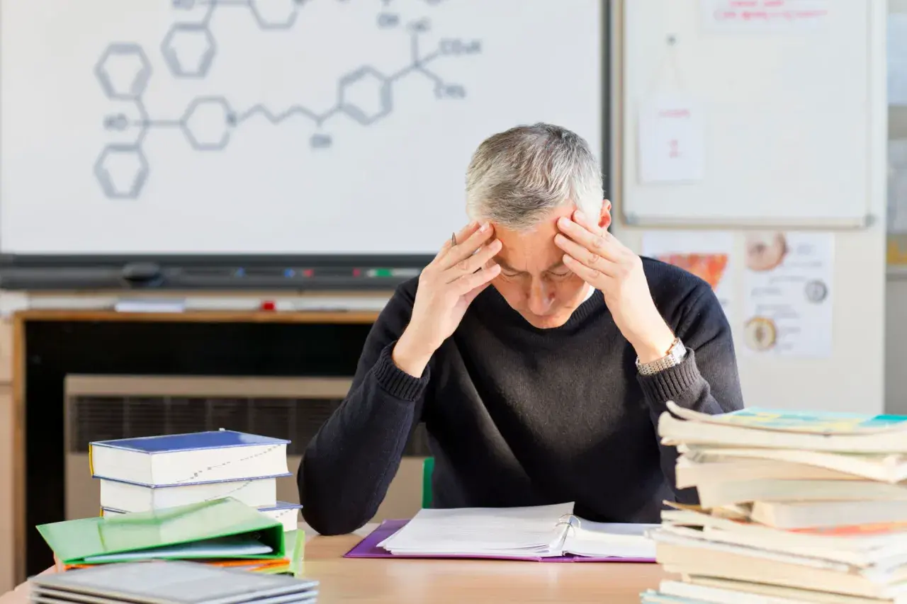 Lehrer mit Händen an den Schläfen, umgeben von Büchern und einer chemischen Formel an der Tafel. Der Lehrer-Stress ist deutlich sichtbar.