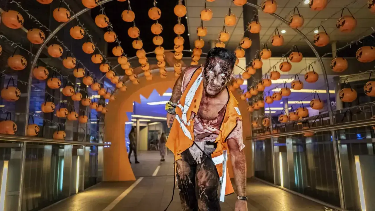 Un espeluznante zombie con maquillaje de Halloween avanza bajo un arco decorado con calabazas en Puerto Venecia.