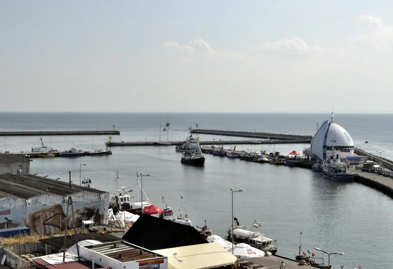 Widok na port Hel z licznymi statkami i łodziami zacumowanymi przy nabrzeżu. W tle widać morze i falochrony.