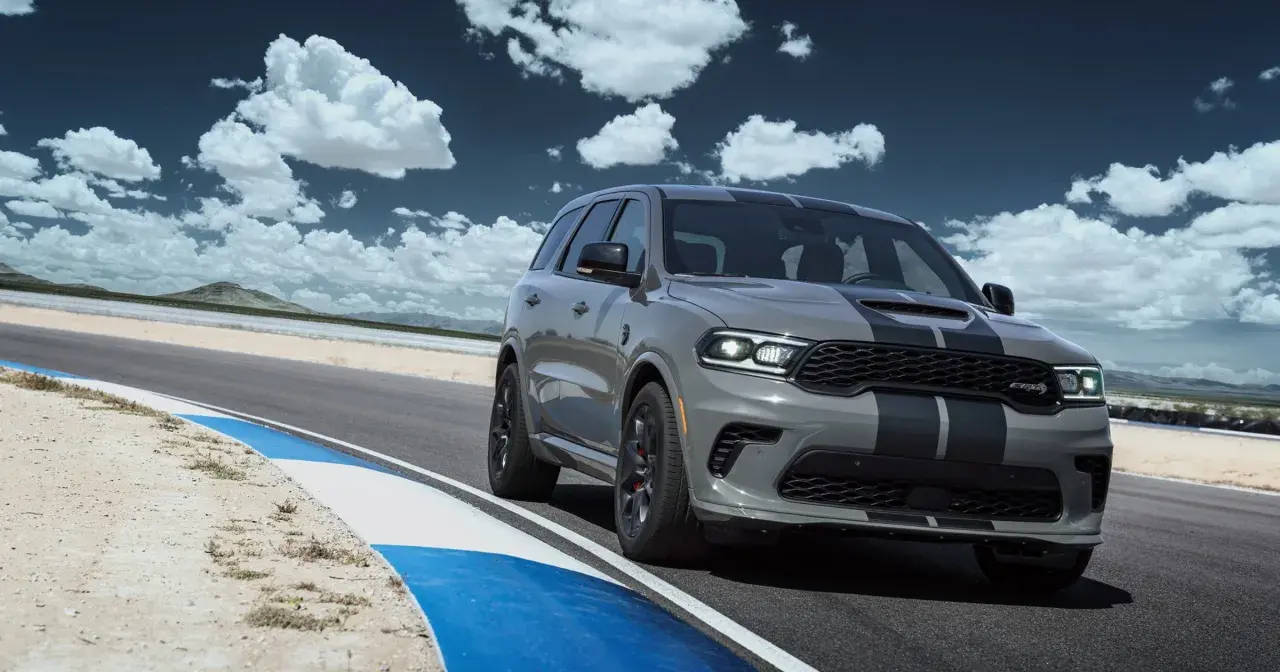Szary Dodge Durango SRT Hellcat z czarnymi pasami pędzi po torze wyścigowym pod błękitnym niebem z chmurami.