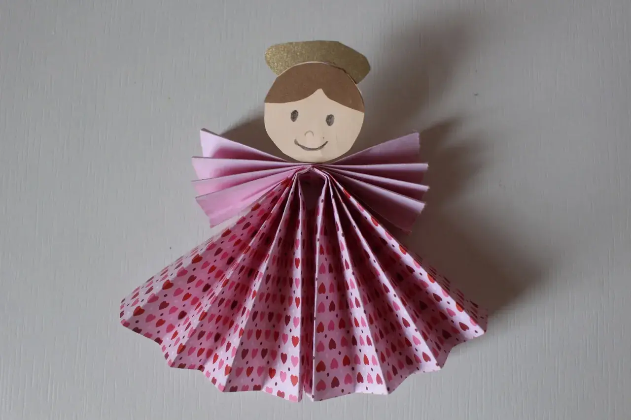 Un adorabile angelo di carta da ritagliare, con un vestito a fantasia di cuoricini rosa e un'aureola dorata.