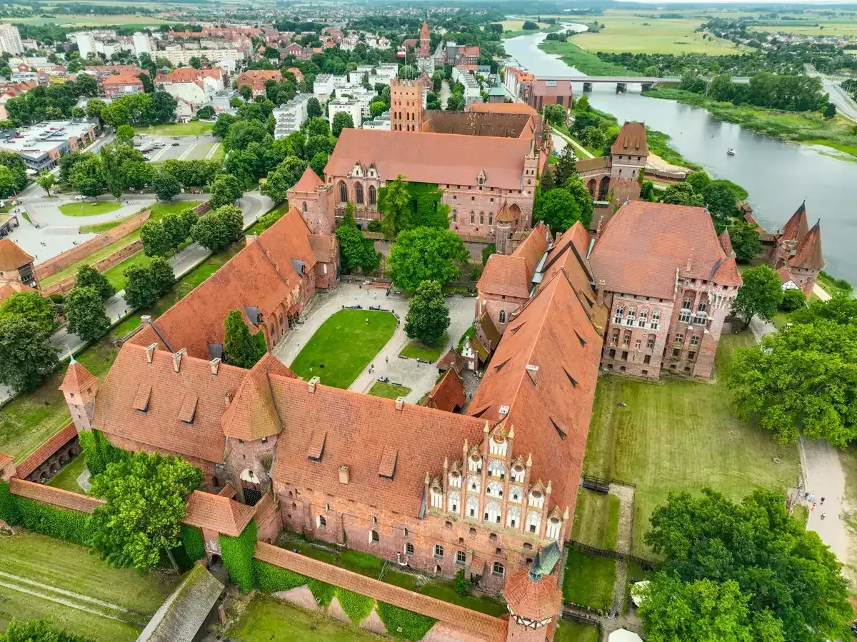 Potężny zamek w Malborku, z czerwonymi dachami i zielonymi dziedzińcami, rozciąga się wzdłuż rzeki Nogat.