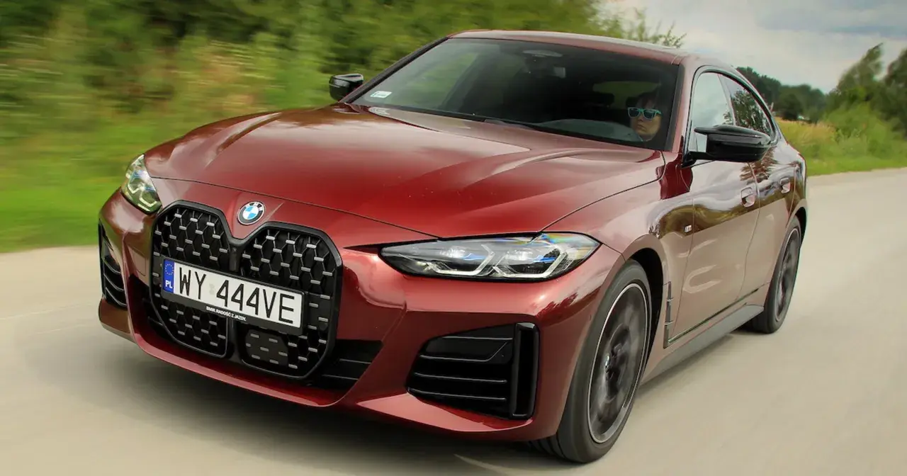 Czerwone BMW M440i z charakterystycznym grillem pędzi po drodze, kierowca w okularach przeciwsłonecznych.