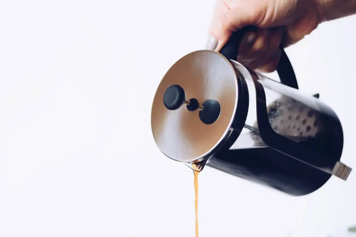 Dłoń trzyma zaparzacz french press, z którego właśnie leje się aromatyczna kawa.