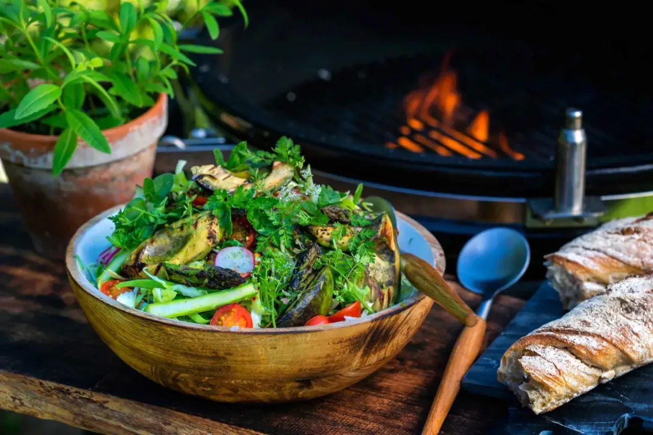 En härlig somrig sallad med grillade grönsaker, tomater och rädisor i en träskål. Bredvid ligger nybakat bröd och en grill med flammor i bakgrunden.