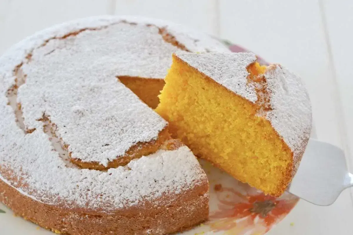 Una fetta di torta di carote e yogurt, soffice e dorata, viene sollevata da una spatola. La torta è cosparsa di zucchero a velo.