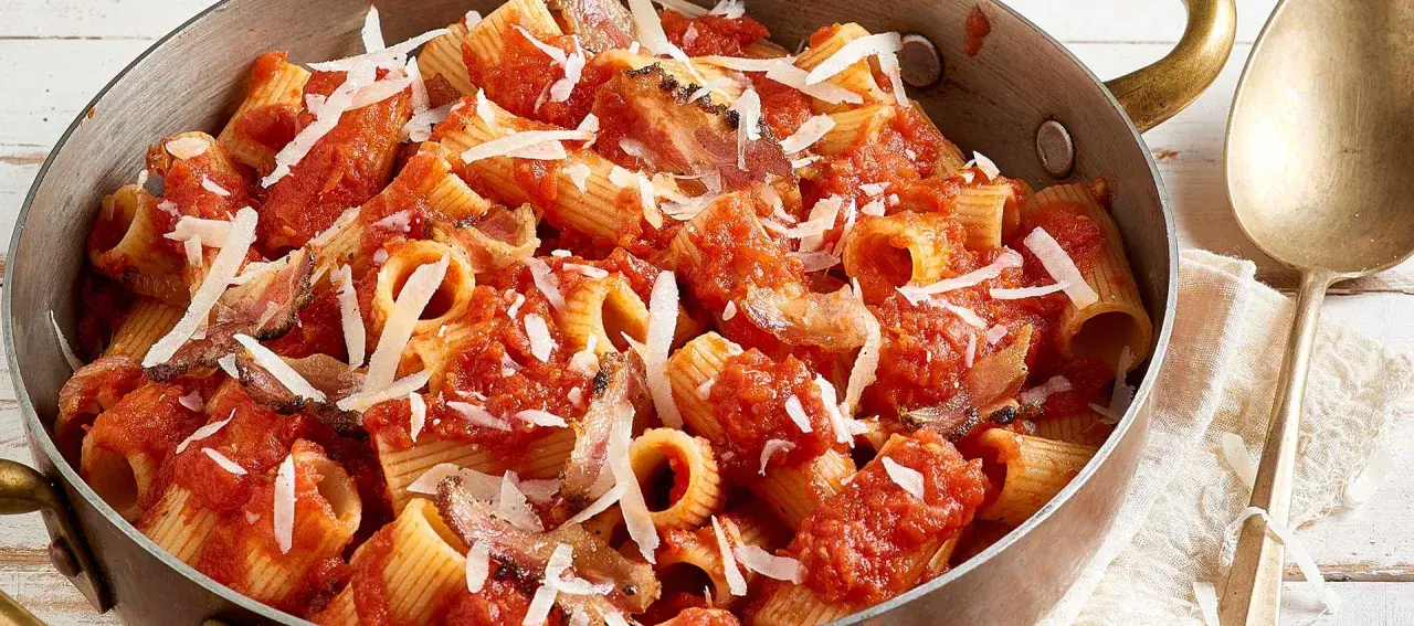 Rigatoni all'amatriciana dans une poêle en cuivre, garni de copeaux de parmesan. Un plat réconfortant et savoureux.