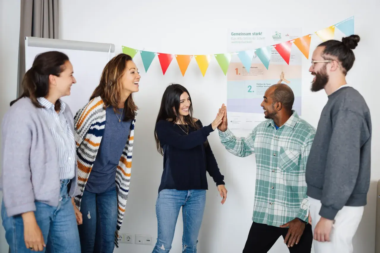 Ein motiviertes Lehrerteam feiert einen Erfolg mit High-Fives. Bunte Wimpelketten und eine Wandtafel deuten auf eine positive Lernumgebung hin.