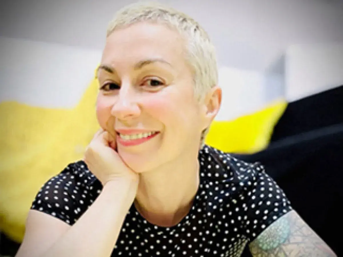 Laura Veltz, a blonde woman with short hair, smiles and rests her chin on her hand. She wears a black polka dot shirt.