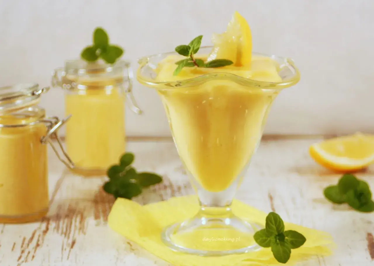 Pyszny, kremowy lemon curd w pucharku, ozdobiony plasterkiem cytryny i miętą. Obok dwa słoiczki z tym samym musem.