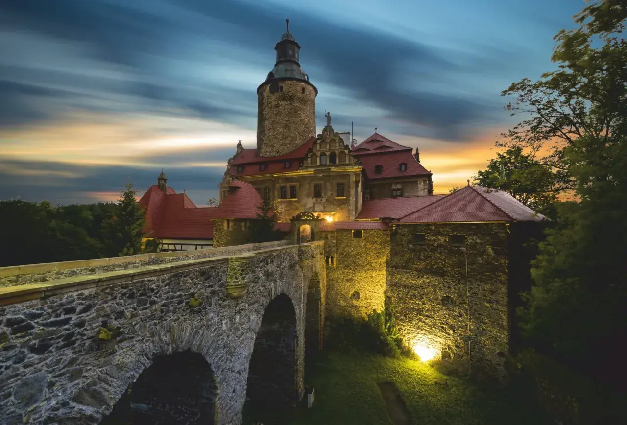 Zamek Czocha, gdzie jest, w blasku zachodzącego słońca i nocnych świateł, z kamiennym mostem i wieżą.